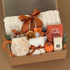 May include: A gift box containing a cream-colored cable knit blanket with a rust-colored ribbon, a ceramic mug with leaf patterns, a dried orange slice, vegan hand cream, a spray bottle, Spicy Pumpkin tea, a pumpkin-shaped candle, and soap.
