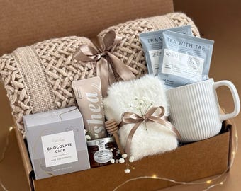 May include: A brown cardboard gift box filled with various items, including a pair of knitted blankets, a mug, a jar of honey, a bag of chocolate chips, a bar of soap, and two tea bags. The box is labeled "Seafoam & Sand" and the tea bags are labeled "Tea with Tae".