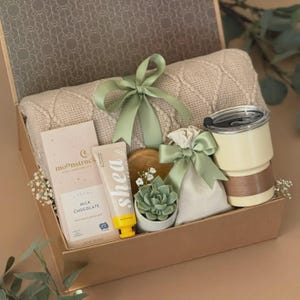 May include: A gift box containing a beige cable-knit blanket tied with a sage green ribbon, a Moonstruck milk chocolate bar, a shea hand cream tube, a small succulent in a white pot, a drawstring pouch, and a cream-colored tumbler with a lid.