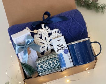 May include: A cozy holiday gift box filled with a blue blanket, a blue enamel mug, coffee, cookies, a macrame snowflake, and socks with a geometric pattern. The box is tied with a blue ribbon and includes a small decorative Christmas tree.