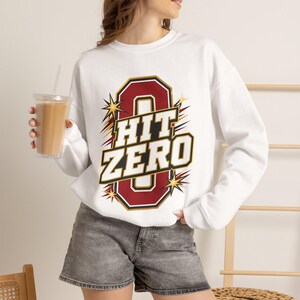 May include: A white long-sleeve sweatshirt featuring a large graphic on the chest. The design includes a bold maroon number zero with a black and gold outline, overlaid with the white text "HIT ZERO" and stylized gold starburst accents.