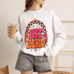 May include: A white long-sleeve sweatshirt featuring a graphic print with the text HIT ZERO in bold pink and orange letters. The design includes a black and white checkered arch, pink lightning bolts, and two yellow smiley faces on the sides.