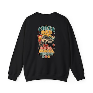 May include: A black long-sleeve sweatshirt featuring a colorful graphic on the back. The design includes a white SUV, a bag of chips, a drink, and the text CHEER DAD RIDES, SNACKS, REPEAT, accented with small stars and red location pin icons.