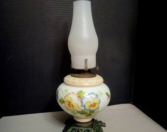May include: A white oil lamp with a green metal base and a white glass shade with a floral design. The floral design features yellow, blue, and pink pansies.
