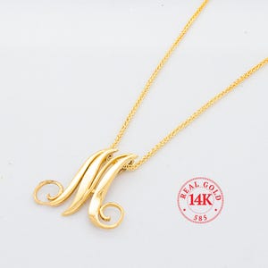 May include: A gold chain necklace with a pendant in the shape of the letter "M". The pendant is made of 14k gold and has a polished finish. The chain is a delicate, rope-style chain.