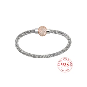 May include: A silver bracelet with a rose gold ball charm encrusted with clear cubic zirconia stones. The bracelet is made of a delicate, textured chain and has a magnetic clasp. The charm is centered on the bracelet and is surrounded by a halo of smaller stones. The bracelet is stamped with "REAL STERLING 925 SILVER".