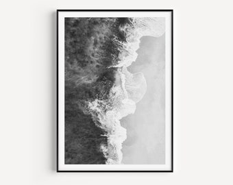 May include: A black and white photograph of ocean waves crashing on a sandy beach. The image is taken from a high angle, looking down at the waves.