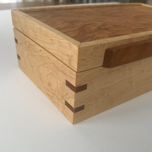 May include: A wooden box with a light-colored exterior and a darker, patterned top. The box features dovetail joints and a brown handle. The wood grain is visible, and the box appears to be handcrafted.