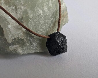 Puede incluir: Un colgante de piedra negra, rugoso y de forma irregular, en un collar de cordón marrón.