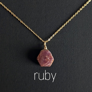 Op de afbeelding: Een gouden ketting met een ruwe robijn hanger. De robijn is diep rood van kleur en heeft een natuurlijke, ruwe textuur. Het woord "robijn" staat in witte tekst onder de hanger.