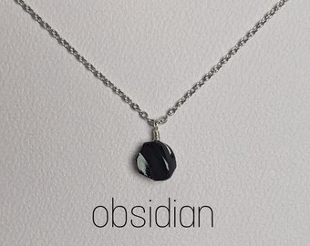 Puede incluir: Un collar de cadena plateada con un colgante de obsidiana negra. El colgante tiene vetas blancas y tiene forma de hexágono redondeado. La palabra "obsidiana" está escrita debajo del colgante.