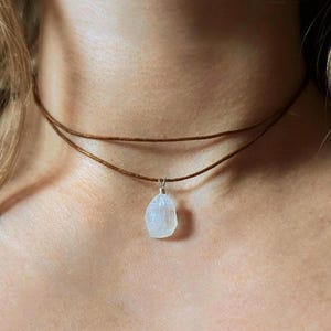 Puede incluir: Un collar gargantilla marrón de doble hebra con un pequeño colgante de color claro de forma irregular. El colgante está suspendido de una fianza de color plateado. El collar se lleva alrededor del cuello.