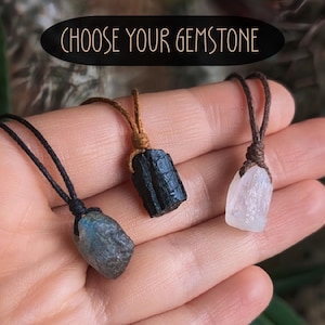 May include: Three gemstone pendants with cord necklaces are shown. The gemstones are a blue-grey, black, and clear crystal. The text "CHOOSE YOUR GEMSTONE" is in a black oval above the pendants.