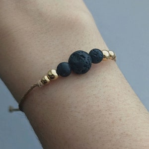 Könnte beinhalten: Ein schlichtes Armband mit schwarzen Lavasteinen und goldenen Perlen auf einer braunen Schnur.