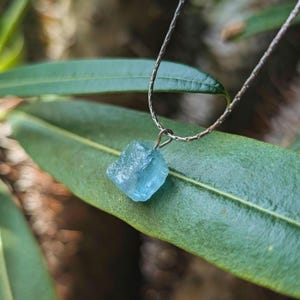 May include: A raw, light blue fluorite crystal pendant on a silver chain. The gemstone is a roughly square shape with a textured surface. The necklace is displayed on a large green leaf, with a blurred background of green foliage.