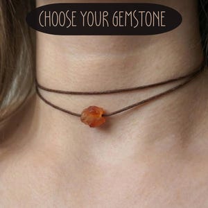 Puede incluir: Un collar gargantilla de doble hebra marrón con un colgante de piedra preciosa naranja en bruto. El collar se lleva alrededor del cuello. El texto "CHOOSE YOUR GEMSTONE" está en un óvalo negro encima del collar.