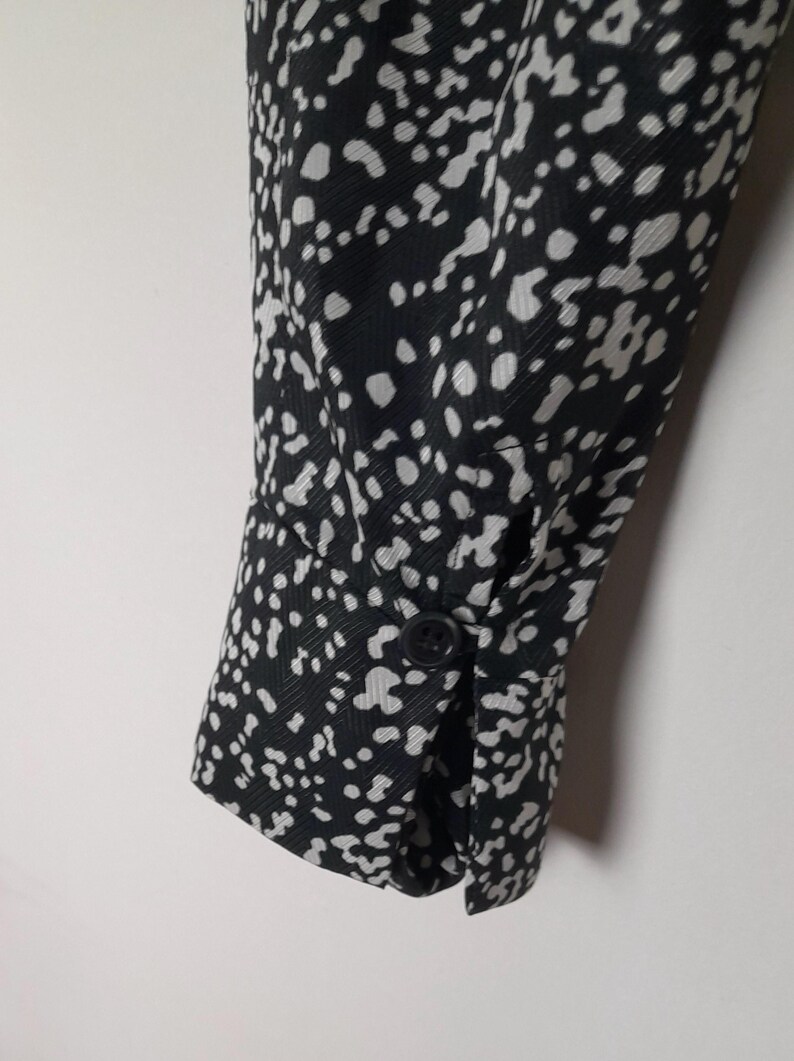 Puede incluir: Primer plano de una manga con estampado en blanco y negro. La tela presenta un patr&oacute;n repetitivo de puntos blancos sobre un fondo negro. El pu&ntilde;o est&aacute; abotonado y la manga est&aacute; ligeramente doblada.