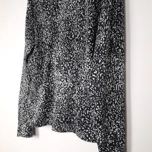 Puede incluir: Una blusa estampada en blanco y negro con mangas largas, colgada de una percha blanca. La tela est&aacute; cubierta de peque&ntilde;os puntos blancos irregulares sobre un fondo negro. La blusa tiene cuello y pu&ntilde;os abotonados.