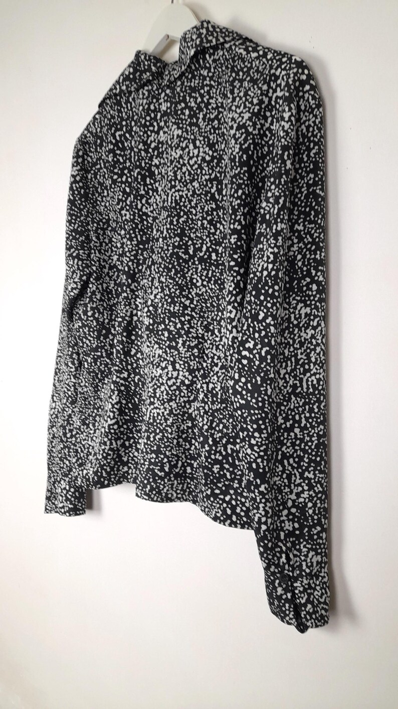 Puede incluir: Una blusa estampada en blanco y negro con mangas largas, colgada de una percha blanca. La tela est&aacute; cubierta de peque&ntilde;os puntos blancos irregulares sobre un fondo negro. La blusa tiene cuello y pu&ntilde;os abotonados.