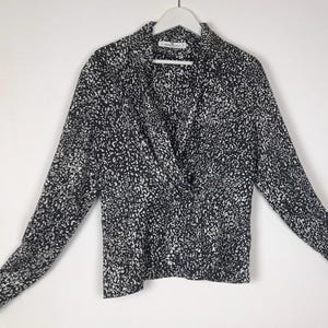 Puede incluir: Una blusa de manga larga con un estampado moteado en blanco y negro. La blusa presenta un frente cruzado y un cuello. Cuelga de una percha blanca sobre un fondo blanco.