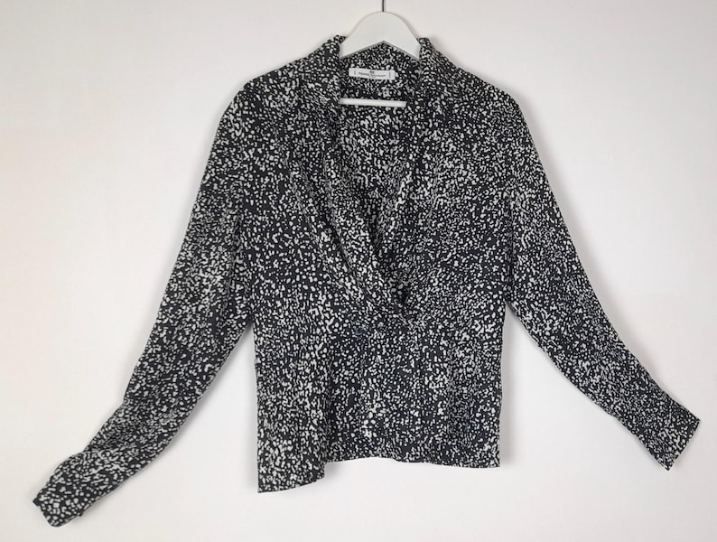 Puede incluir: Una blusa de manga larga con un estampado moteado en blanco y negro. La blusa presenta un frente cruzado y un cuello. Cuelga de una percha blanca sobre un fondo blanco.