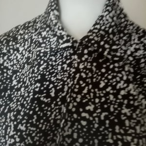 Puede incluir: Una camisa con cuello con estampado en blanco y negro. La tela presenta un dise&ntilde;o moteado con peque&ntilde;os puntos blancos sobre un fondo negro. La camisa est&aacute; abotonada en el cuello y parece estar hecha de un material ligero.