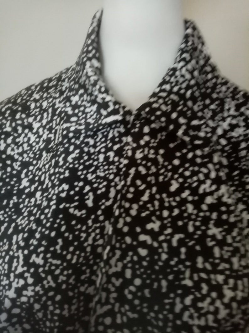 Puede incluir: Una camisa con cuello con estampado en blanco y negro. La tela presenta un dise&ntilde;o moteado con peque&ntilde;os puntos blancos sobre un fondo negro. La camisa est&aacute; abotonada en el cuello y parece estar hecha de un material ligero.