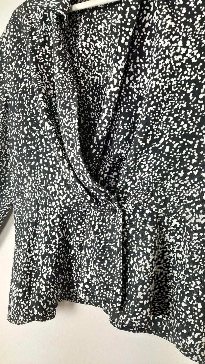 Puede incluir: Top con estampado en blanco y negro con cuello drapeado y cierre de botones. La tela presenta un dise&ntilde;o moteado, creando una apariencia texturizada. El top tiene mangas largas y un ajuste entallado, adecuado para diversas ocasiones.