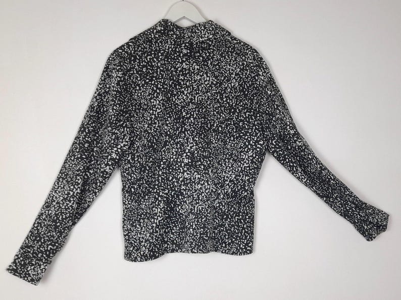 Puede incluir: Una camisa de manga larga con estampado en blanco y negro. La camisa est&aacute; colgada de una percha blanca sobre un fondo blanco. El estampado es un dise&ntilde;o moteado, con peque&ntilde;os puntos blancos sobre una base negra.