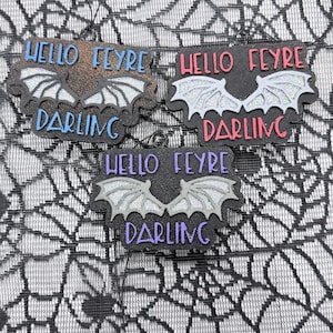 May include: Three black hanging ornaments featuring the text HELLO FEYRE DARLING and a pair of glittery silver bat wings. One ornament has blue text, one has red text, and one has purple text. Each piece includes a small black hanging loop.
