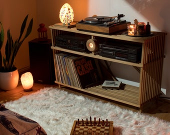 May include: A wooden shelf unit with a record player, a stack of vinyl records, a chessboard, and two lamps. The shelf unit is made of light wood and has a rustic look. The record player is black and has a silver turntable. The vinyl records are stacked on the shelf and are labeled with the name "Johnny Cash". The chessboard is made of wood and has black and white squares. The lamps are both on and are casting a warm glow on the shelf unit.