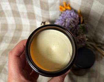 May include: A top-down view of an open amber glass jar containing a smooth, pale yellow cream. The jar has a black rim and is held in place, with a second closed amber jar and a small cluster of dried purple and orange flowers visible nearby.