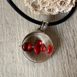 Puede incluir: Un collar con un colgante redondo de plata y cuentas de coral rojo. El collar está en un cordón negro.