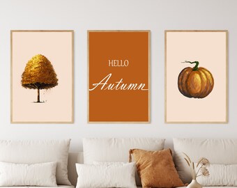 May include: Three framed prints with an autumn theme. The left print features a yellow tree on a cream background. The center print has an orange background with the text "HELLO AUTUMN" in white. The right print features a brown pumpkin on a cream background.