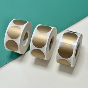 May include: Three rolls of gold round stickers. Each roll has a white backing and is partially unwound, revealing the gold stickers.