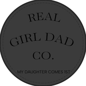 May include: A circular dark grey patch featuring the black text REAL GIRL DAD CO. in the centre and the text MY DAUGHTER COMES 1ST along the bottom edge. The circular design includes a thin stitched border detail around the outer perimeter.