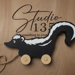 May include: A wooden pull toy skunk with black and white markings, rolling on two wooden wheels. The toy has a rope attached for pulling. The background is a light wood surface with the text "Studio 135" in a script font.