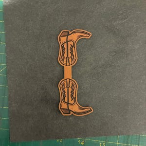 May include: A laser-cut, tan-colored cowboy boot design. The boot features intricate detailing and is connected at the top and bottom. The design is set against a dark gray background with a ruler along the bottom edge.