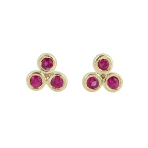 May include: A pair of gold stud earrings, each featuring three round, vibrant pink gemstones arranged in a triangular cluster. The stones are set in smooth, polished gold bezels that connect to form a cohesive, three-part floral-like shape.