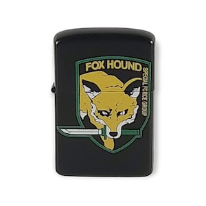 Puede incluir: Encendedor negro con un emblema Fox Hound verde, amarillo y blanco. El emblema presenta un zorro sosteniendo un cuchillo en la boca, con el texto "FOX HOUND" y "SPECIAL FORCE GROUP".
