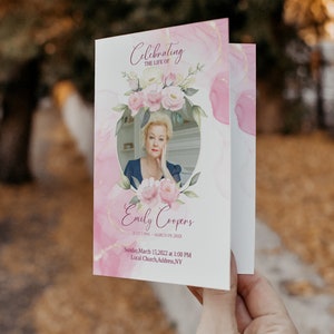 May include: A pink and white funeral program with a photo of a woman in a floral wreath. The text reads "Celebrating the Life of Emily Coopers July 7 1945 - March 01 20XX Sunday, March 15, 2022 at 1:00 PM Local Church, Address, NY"