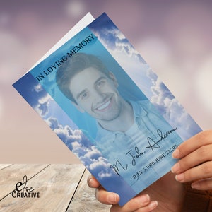 Peut inclure: Une carte de deuil avec un ciel bleu et des nuages, avec la photo d'une personne souriante. Le texte indique "IN LOVING MEMORY" et "M. John Anderson JULY 5, 1970-JUNE 22, 203". La carte est tenue dans une main.