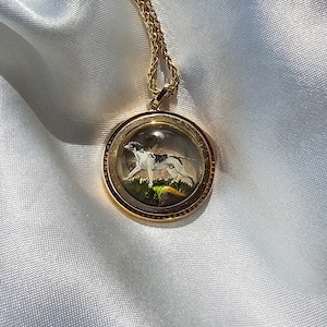 May include: A gold-toned pendant necklace featuring a round, clear glass charm with a painted image of a dog. The dog is white with black spots, standing on green grass. The necklace chain is also gold-toned.