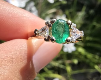 May include: A gold ring featuring a large oval-cut emerald center stone, flanked by two smaller, triangular-cut diamonds. The ring is held up against a blurred background of green foliage and white flowers.