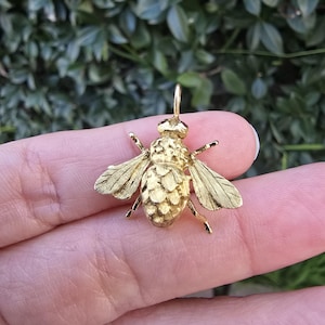 May include: A gold-colored bee pendant with detailed wings and body. The pendant has a small loop at the top for a chain. The bee's body has a textured, segmented design. The wings are delicately etched.