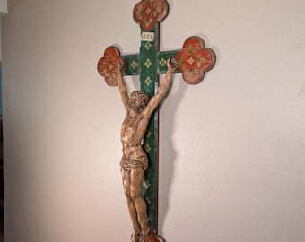 May include: A large wooden crucifix featuring a carved, light-brown figure of Jesus mounted on a dark green cross. The cross has trefoil-shaped ends painted in red with floral patterns and includes the white text INRI at the top.