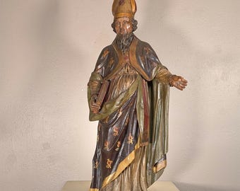 May include: A carved wooden statue of a bearded bishop wearing a tall miter and layered robes in shades of dark brown, gold, and muted green. The figure holds a book in one hand and extends the other, featuring intricate painted floral details.