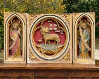 May include: A triptych featuring three carved panels. The side panels depict angels in robes holding scrolls. The central circular panel shows a lamb standing on a mound, holding a cross-topped staff with a flag, positioned above a chalice.