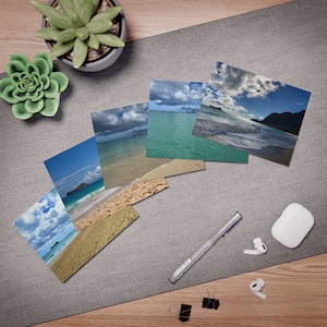 May include: A collection of five rectangular photographic prints featuring tropical beach scenes with turquoise water, sandy shores, and cloudy blue skies, arranged in a fan shape alongside a gray pen, white wireless earbuds, and black clips.