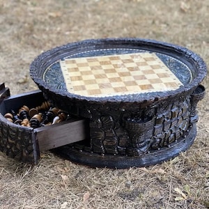 May include: A wooden chess table with a carved design. The table has a round top with a chessboard inlay and a drawer that pulls out to reveal chess pieces.